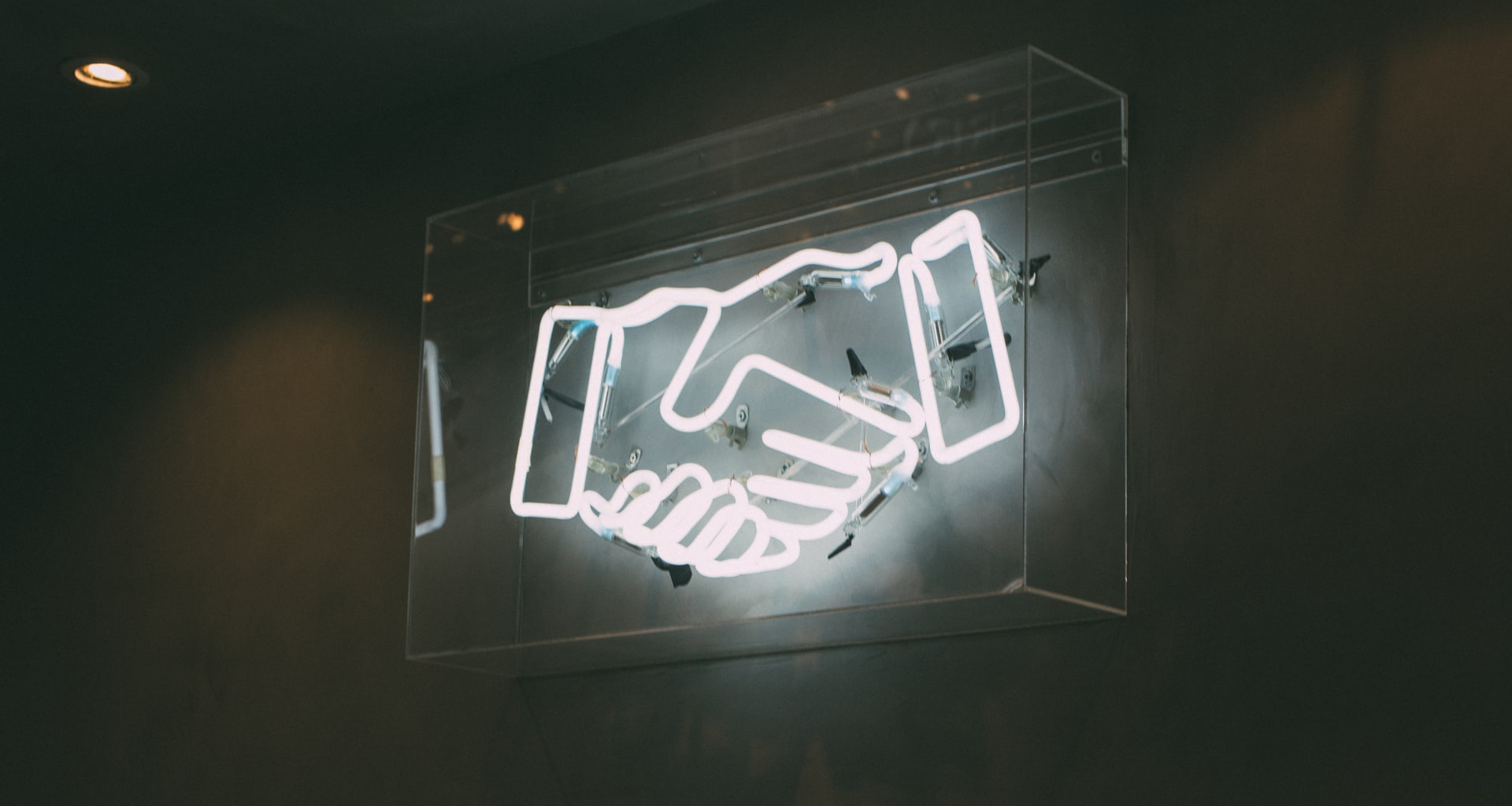 A white neon light sign of two hands shaking, encased in a glass box, hanging on a dark grey wall