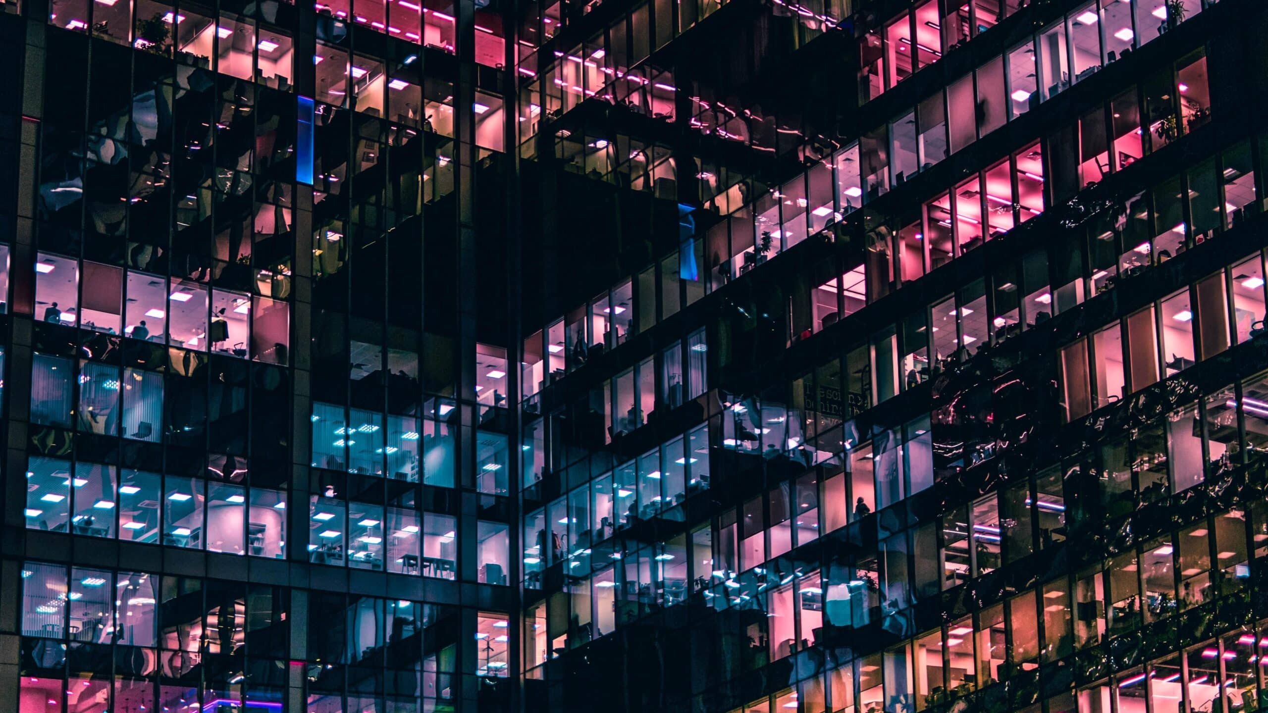 A ground view looking upwards at a lot of different office spaces, high up in an apartment building, with pink and blue lighting