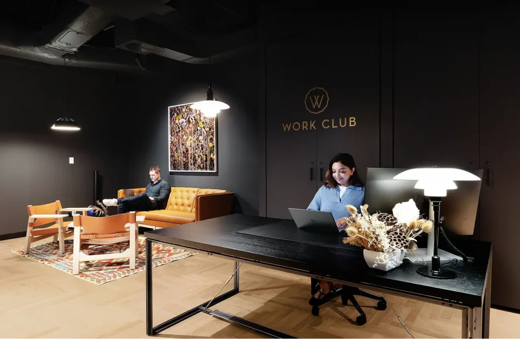 A woman types on a laptop at a black desk while a man works on a sofa nearby inside a dark lounge featuring a gold wall emblem with a "W" above text reading "WORK CLUB".