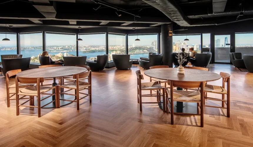 Round wooden dining tables paired with woven chairs sit on a herringbone floor in a high-rise lounge, where panoramic windows offer sweeping views of a city harbor and bridge.