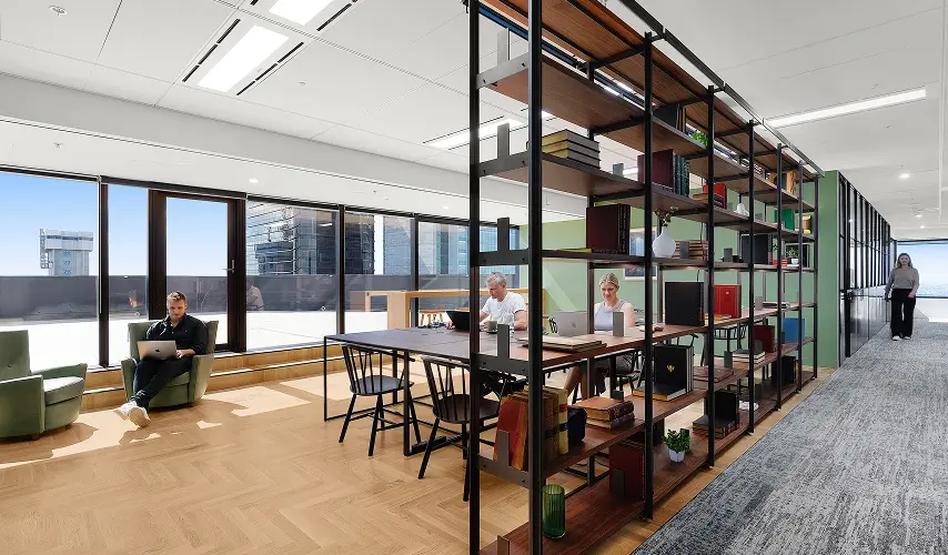 People work on laptops at tables and in armchairs inside a modern, sunlit office space featuring large floor-to-ceiling windows and a tall metal shelving divider filled with books.