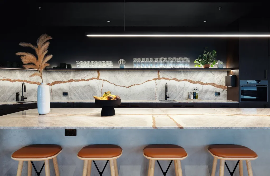 Four brown leather stools sit beneath a large marble island in a sleek dark kitchen with an illuminated veined backsplash, floating shelves holding glassware, and a linear pendant light.