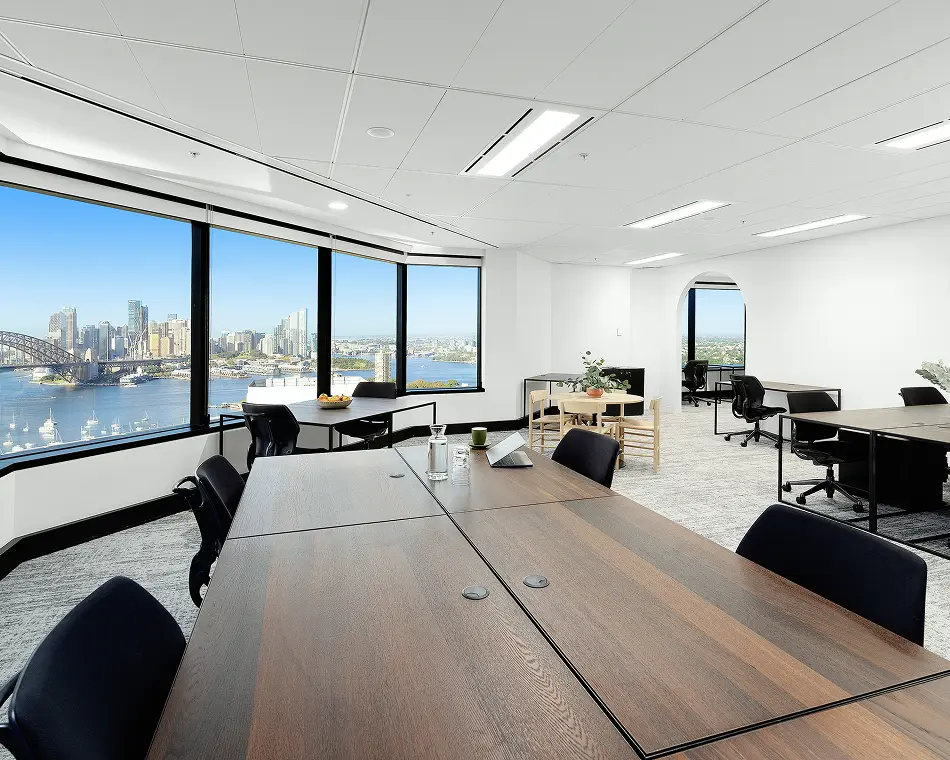 Dark wooden conference tables paired with black office chairs sit in a bright, modern open-plan office space featuring panoramic floor-to-ceiling windows overlooking a sunny harbor and city skyline.