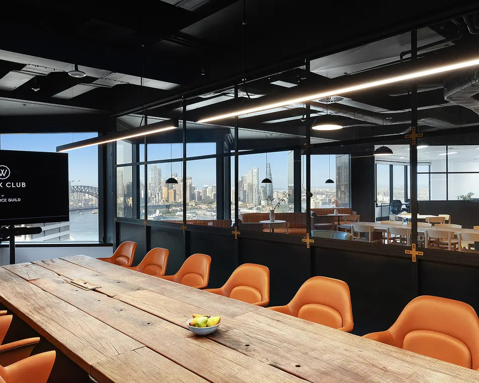 Bright orange armchairs surround a long, rustic wooden conference table inside a high-rise office suite featuring glass partition walls that overlook a panoramic city skyline. A monitor on the left displays the text: "(W) K CLUB BY ICE GUILD"