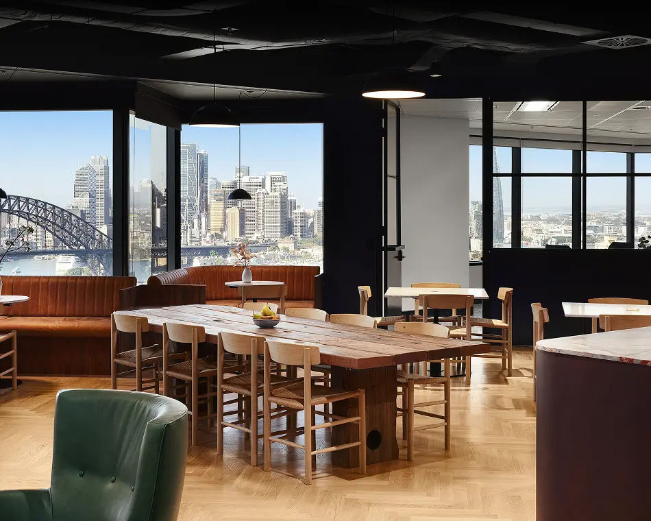 A long wooden dining table surrounded by light wood chairs sits centrally within a modern office lounge featuring parquet flooring and large windows overlooking a city skyline and harbor bridge.