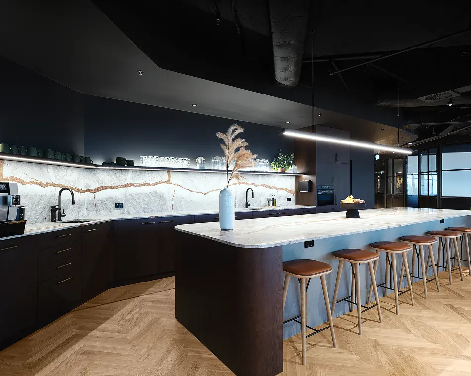 A long marble-topped kitchen island lined with wooden bar stools anchors a modern space featuring dark cabinetry, a backlit marble backsplash, linear lighting, and herringbone wood floors.
