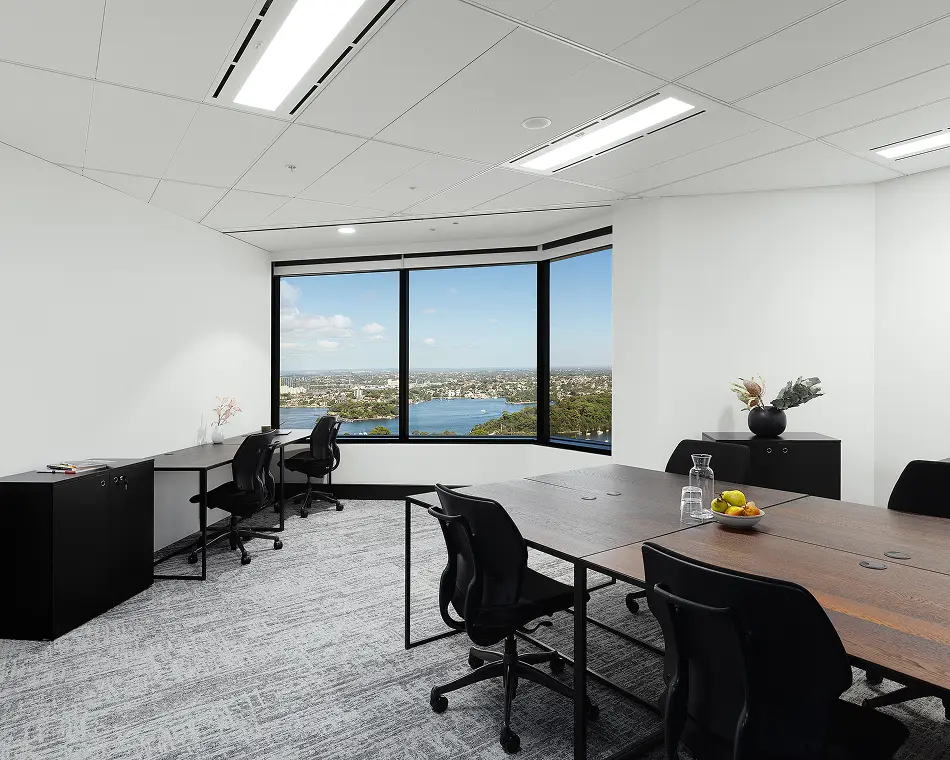 Dark wooden desks and black rolling chairs furnish a modern, bright office room, set against panoramic windows overlooking a blue bay and cityscape under a clear sky.