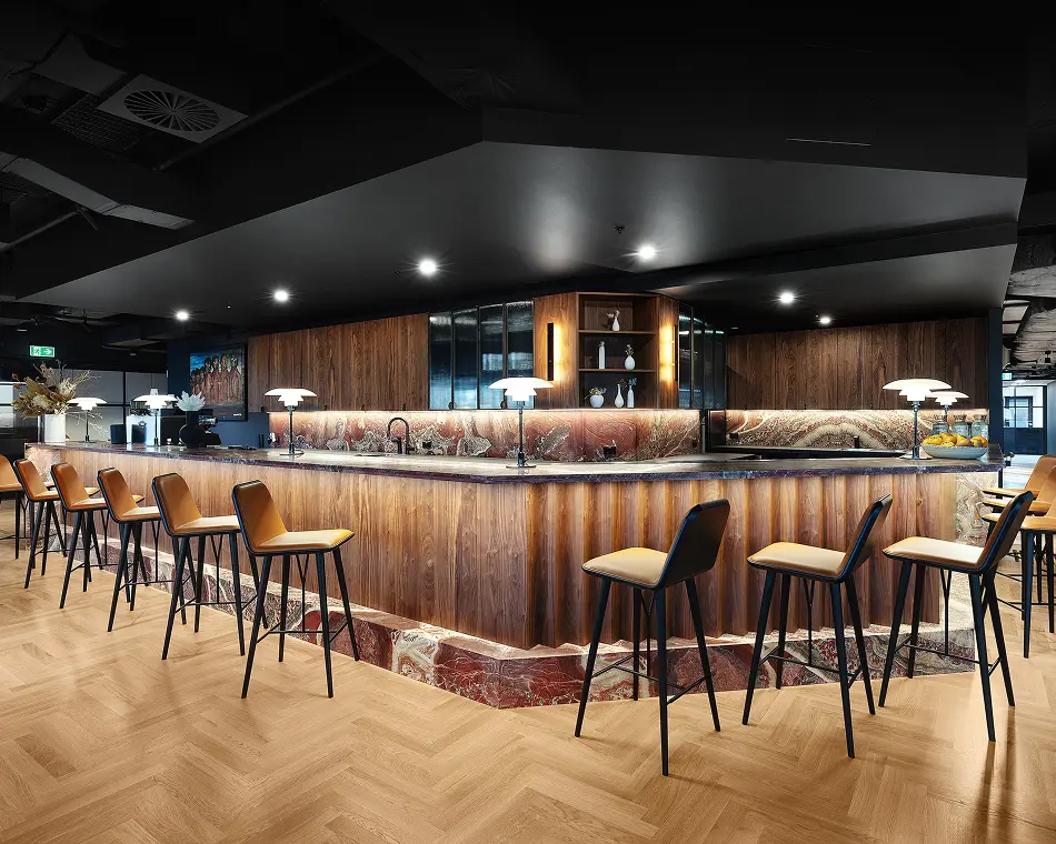 A curved, wood-paneled bar counter lined with tan leather stools glows under warm lamps, set on a light herringbone wood floor beneath a dark ceiling inside a modern lounge.