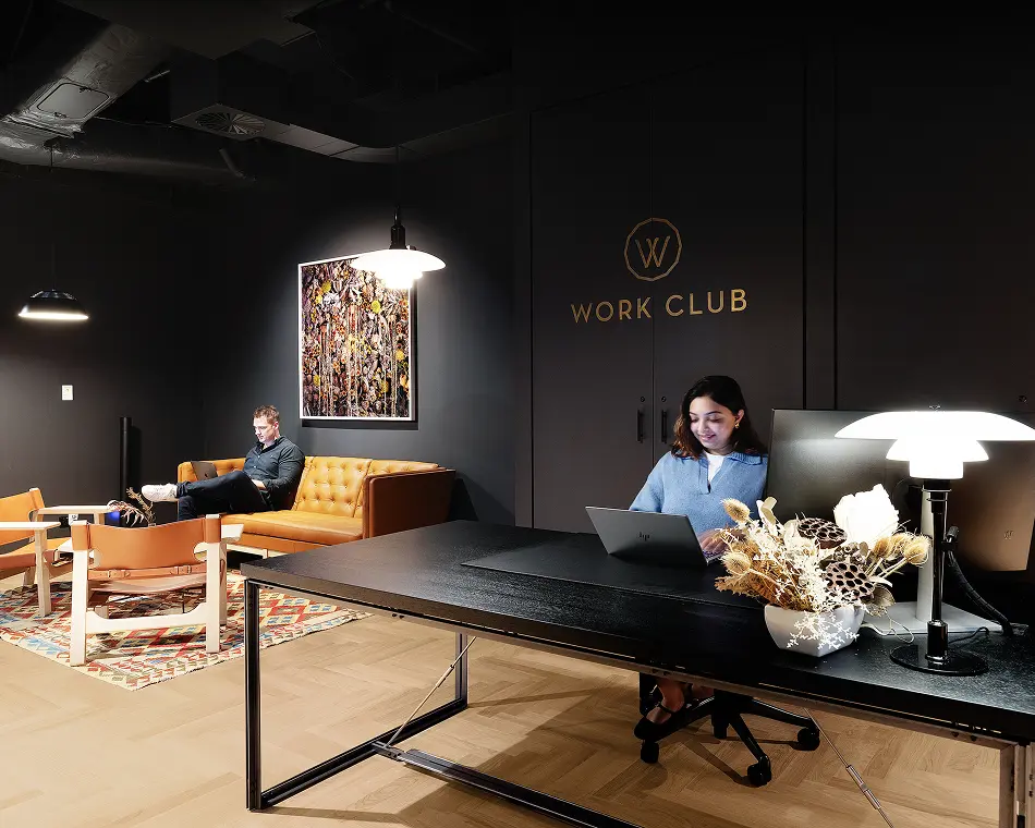 A woman works at a dark desk while a man uses a laptop on a leather couch inside a dimly lit co-working lounge featuring wall text that reads “WORK CLUB”.
