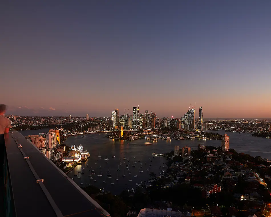 An illuminated city skyline and prominent steel arch bridge glow across a calm harbor at dusk, viewed from a high rooftop terrace overlooking boats anchored in the water below.
