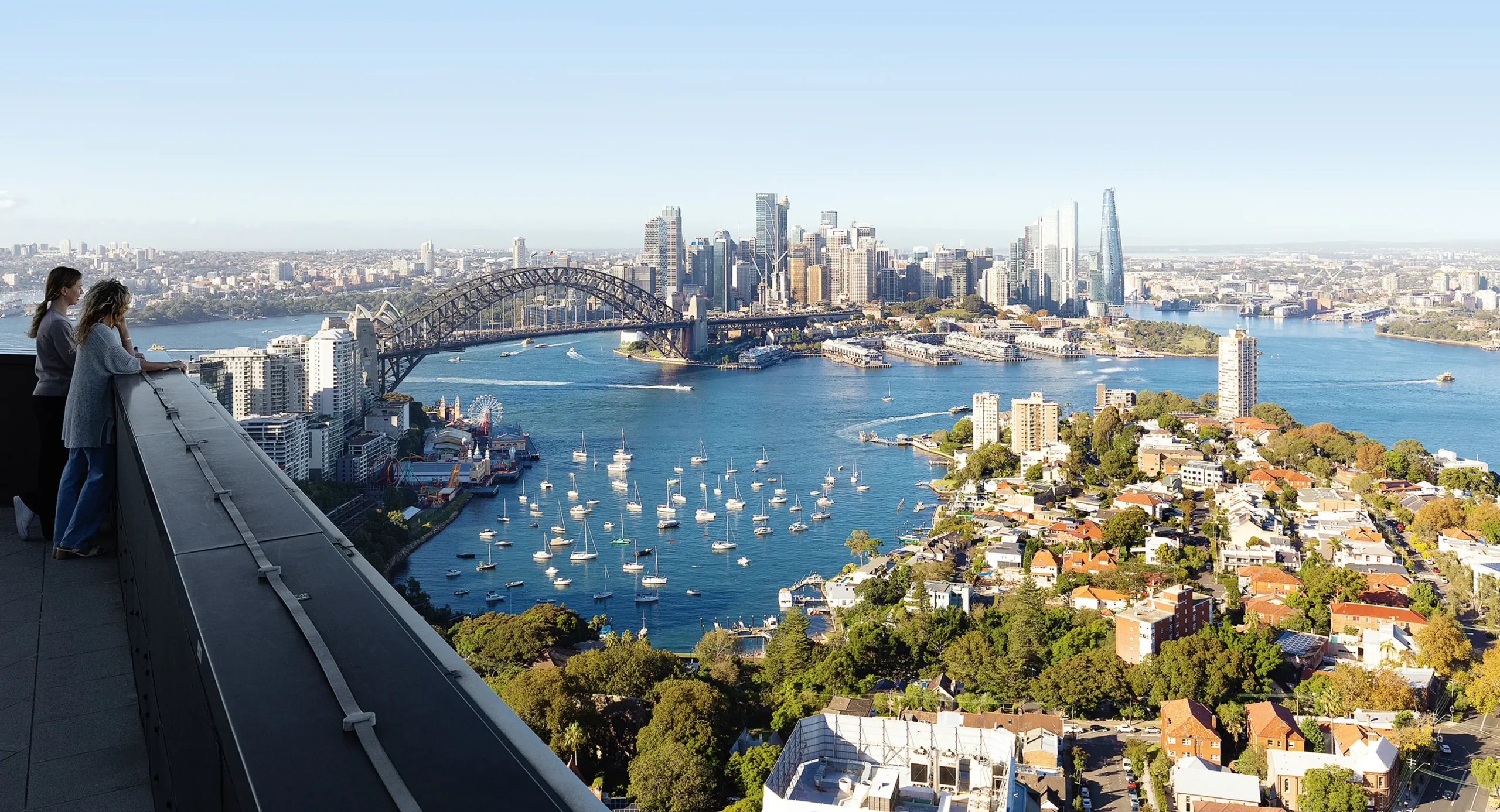 Two women lean on a high-rise balcony ledge, looking out at a panoramic view of Sydney Harbour, featuring the Harbour Bridge, boats, and the distant city skyline.