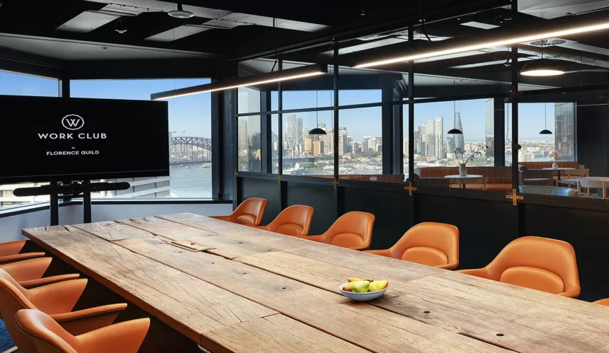 A long wooden conference table surrounded by orange armchairs sits inside a modern high-rise boardroom overlooking a city harbor through large windows. A screen on the left displays text: "W WORK CLUB BY FLORENCE GUILD".
