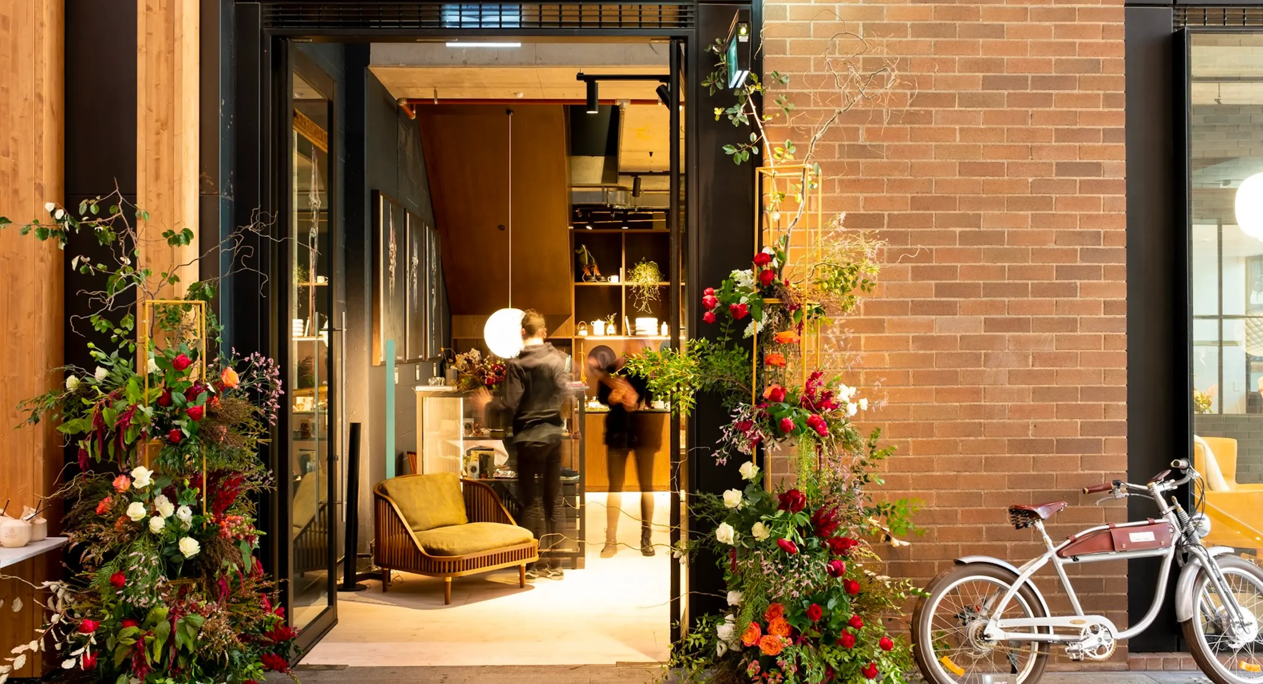 Vibrant floral arrangements frame the open entrance of a modern shop as figures move inside. The doorway is set along a brick wall with a vintage bicycle parked nearby.