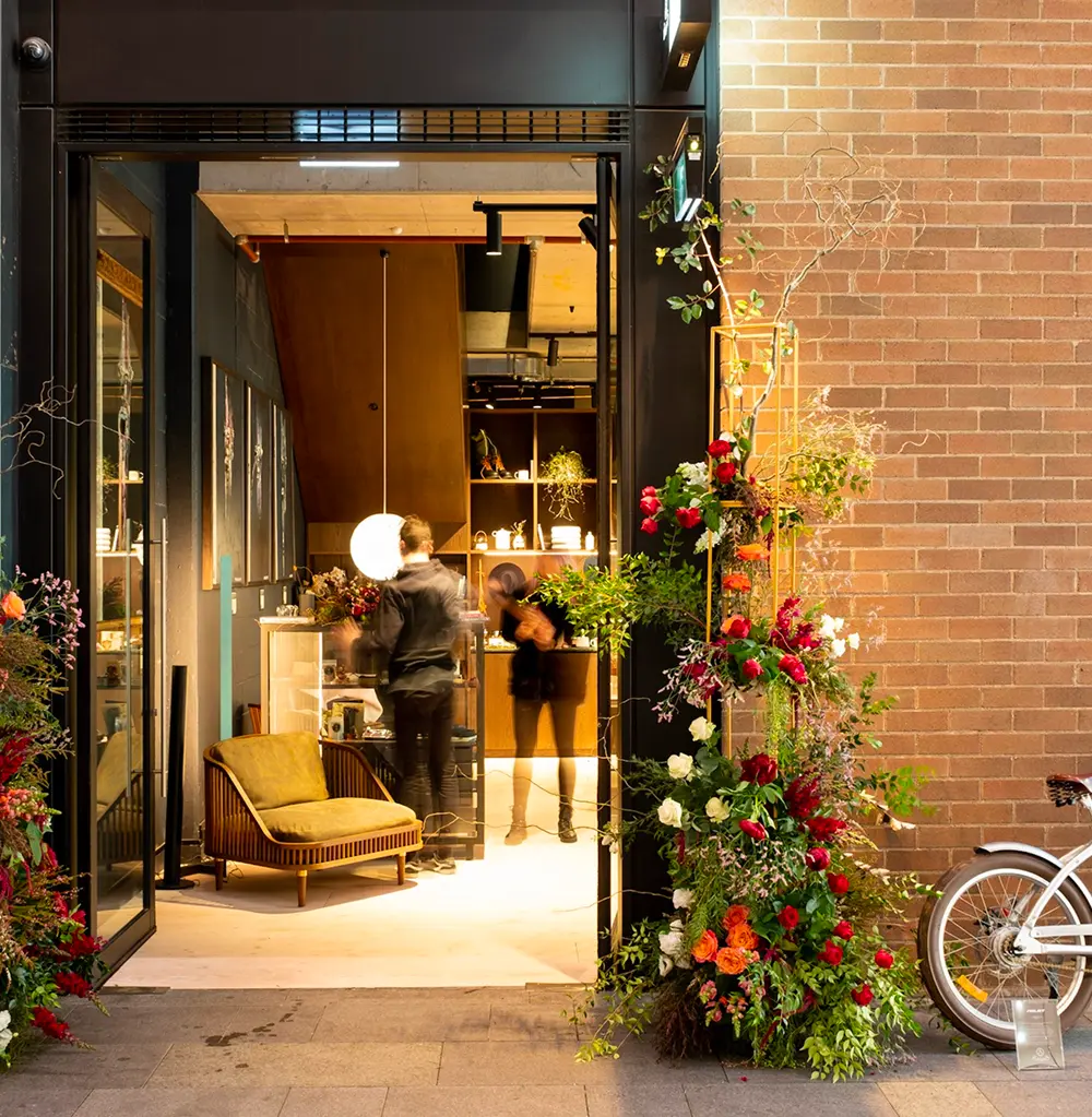 Two figures move inside a warm, dimly lit boutique seen through an open doorway framed by tall floral arrangements, positioned next to a brick wall and a parked bicycle.