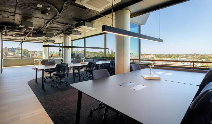 Modern grey office desks paired with ergonomic chairs sit arranged beneath linear pendant lights in a spacious room featuring exposed ceiling pipes and wall-to-wall windows overlooking a sunny cityscape.