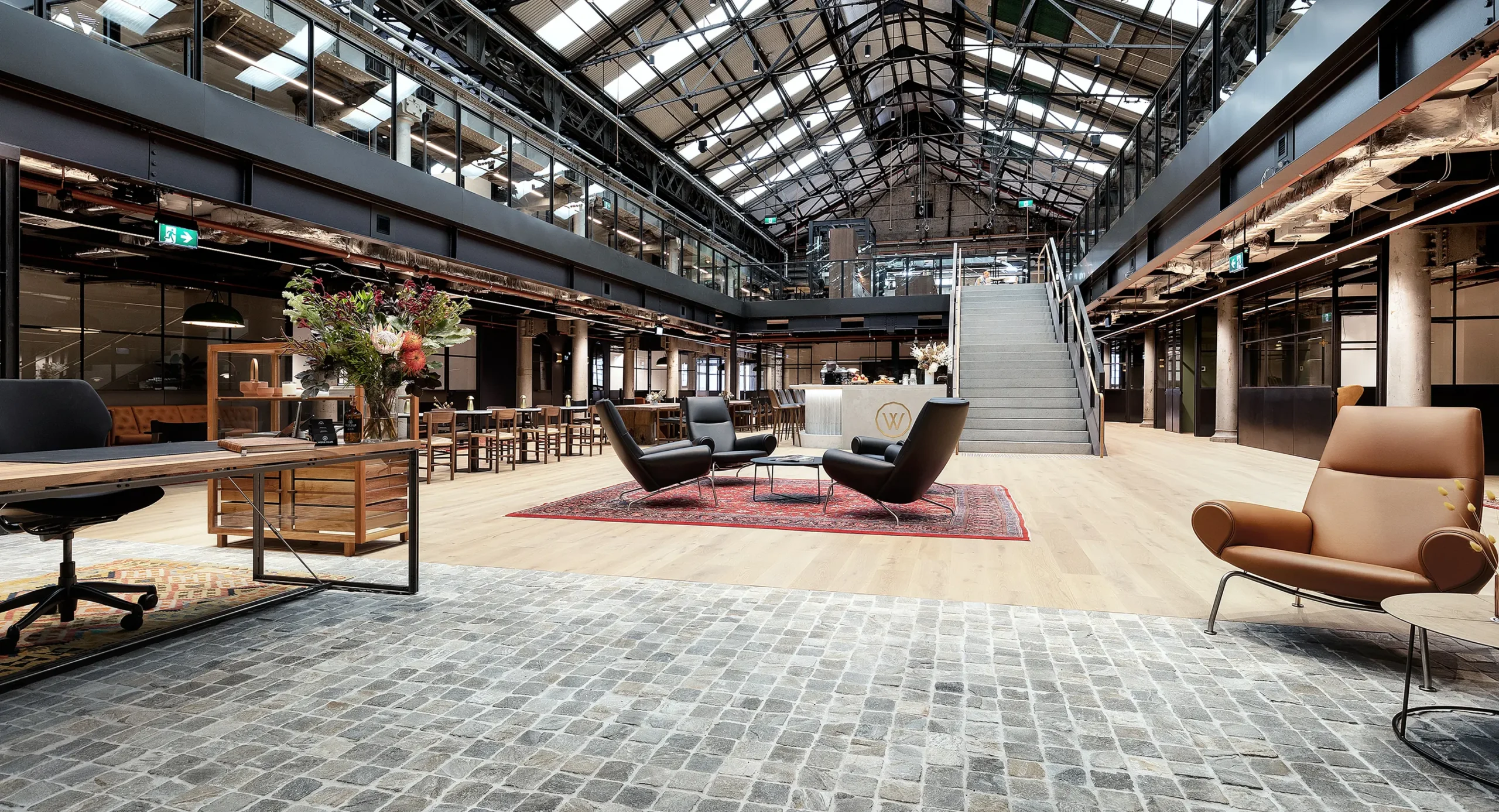 Black leather armchairs sit around a red patterned rug inside a spacious industrial-style office featuring light wooden floors, a central staircase, mezzanine levels, and a vaulted glass skylight ceiling. Text reads: "WORK CLUB", "W".