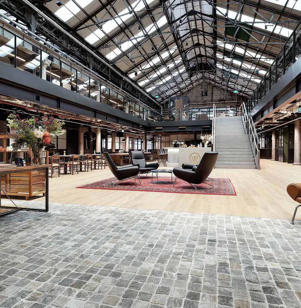 Black leather lounge chairs surround a small table on a red rug within a multi-level industrial atrium featuring a high vaulted skylight ceiling, wooden floors, and a wide staircase. A circular logo containing the letter "W" is visible on the counter in the background.
