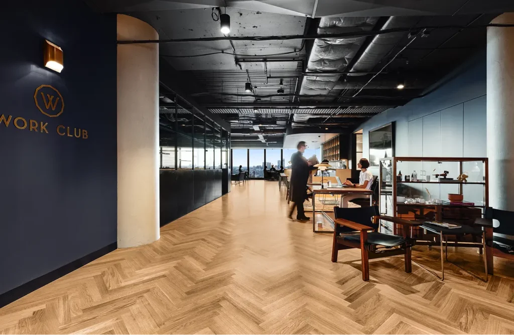 People walk and work inside a modern office featuring herringbone wood flooring and an exposed ceiling, beside a dark wall reading "W WORK CLUB".