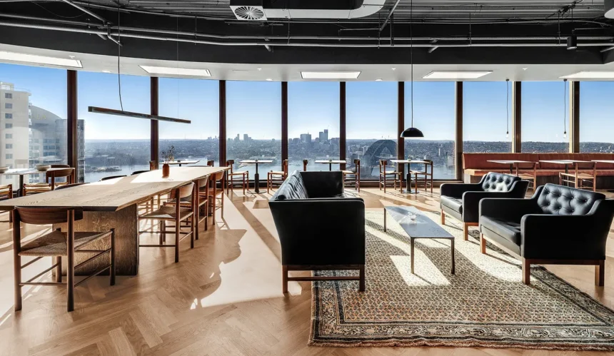 Modern black leather seating and a long wooden table furnish a spacious high-rise lounge, offering panoramic views of the Sydney Harbour Bridge through expansive floor-to-ceiling windows.