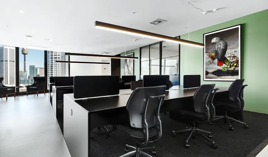 Modern black desks paired with ergonomic chairs sit arranged under linear strip lights inside a contemporary office space featuring a green accent wall with framed artwork and city skyline views.