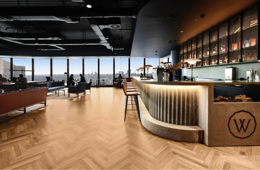 A stylish curved bar counter with warm under-lighting and barstools stands in a spacious high-rise lounge with herringbone wooden floors, seating, and floor-to-ceiling windows overlooking a city skyline. Text visible in the image includes: "W" (inside an octagonal logo on the front of the bar), "...i-LA" (on a distant building through the far-left window).