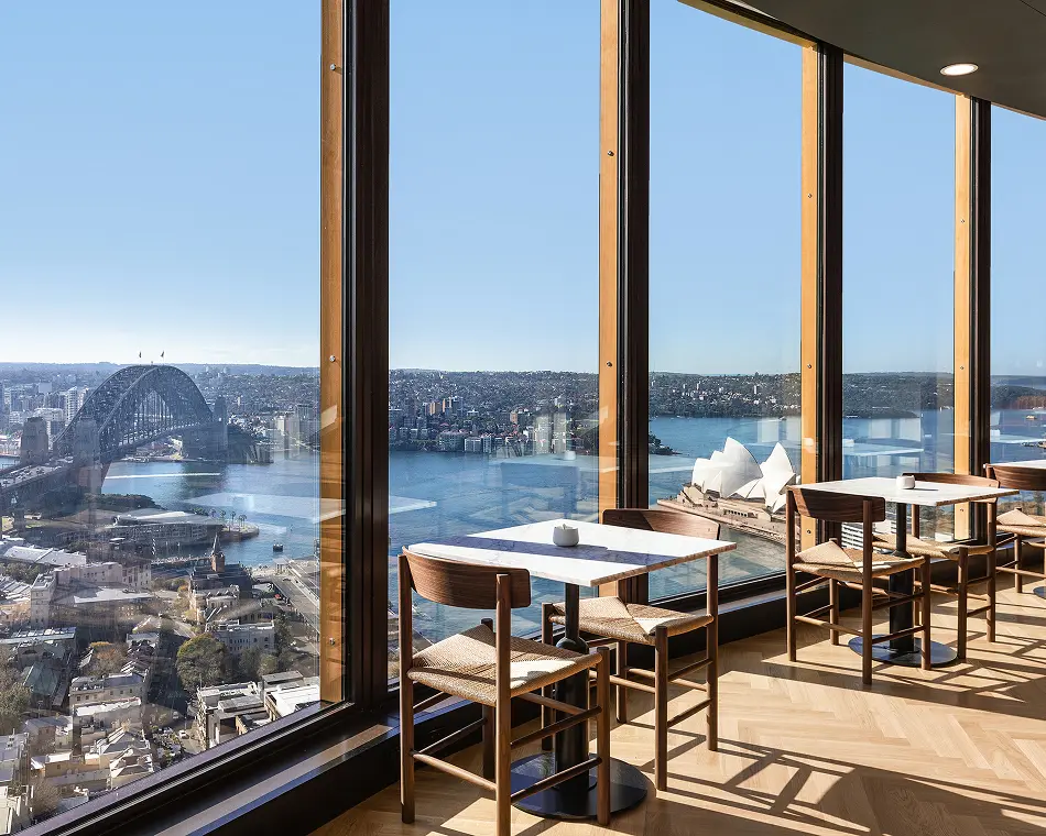 Modern dining tables and wooden chairs line floor-to-ceiling glass windows, offering a panoramic view of the Sydney Harbour Bridge, Sydney Opera House, and sunny harbor below.