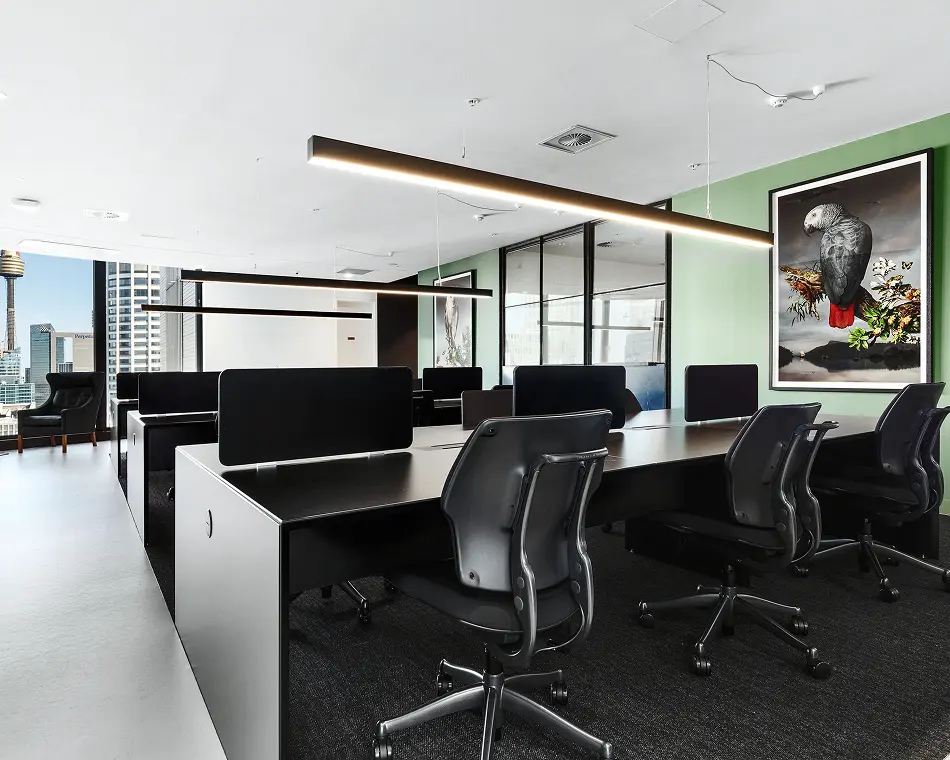 Black office workstations paired with ergonomic chairs sit arranged in rows beneath linear hanging lights, set within a modern office featuring light-green walls, artwork, and high-rise city views.
