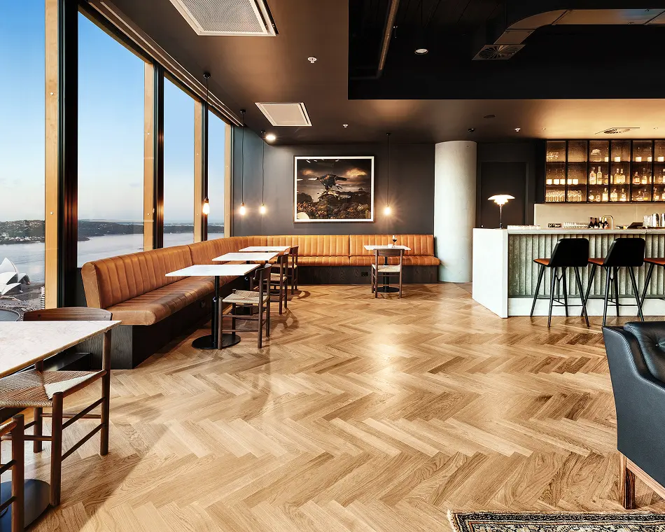 A modern upscale lounge, featuring tan leather banquettes, herringbone wood floors, and small dining tables, lines floor-to-ceiling windows that offer panoramic views of a harbor and city skyline.