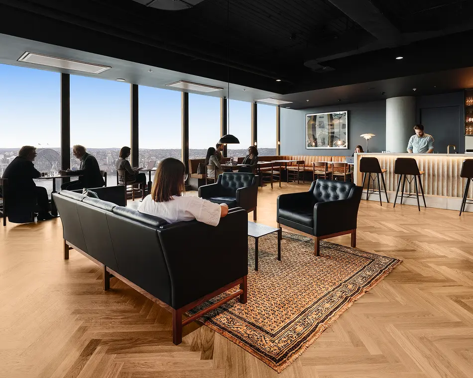 People sit on sleek black leather furniture and at window-side tables, conversing and working inside a modern high-rise lounge featuring herringbone parquet flooring, a bar, and expansive city harbor views.