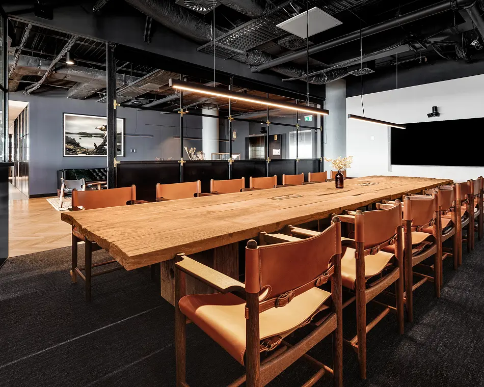 A long, rustic wooden conference table surrounded by tan leather chairs sits in a modern boardroom, illuminated by linear pendant lights beneath an exposed, dark industrial ceiling.