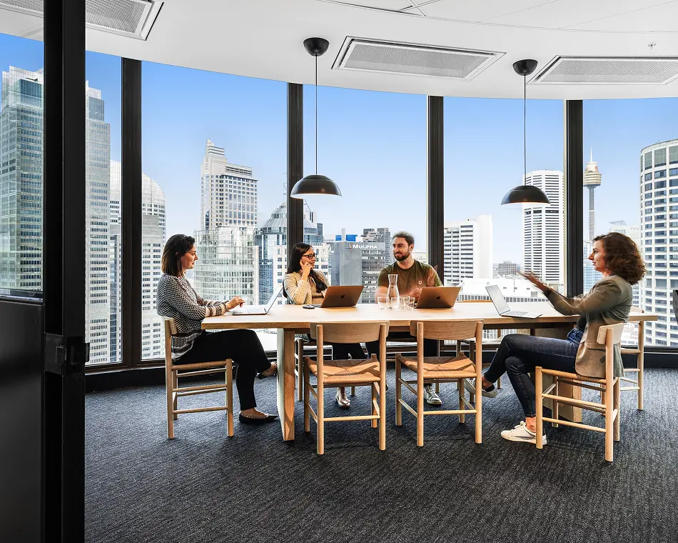 Four colleagues sit around a wooden conference table, conversing and using laptops inside a high-rise office with panoramic windows overlooking a city skyline. Text on a background building reads: "MULPHA".