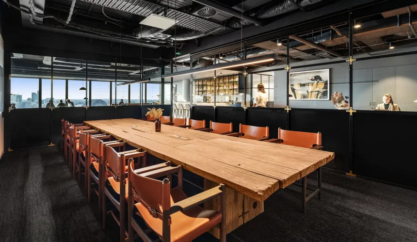 A long, rustic wooden conference table flanked by tan leather chairs sits in a modern office enclosed by dark glass partitions, black industrial ceilings, and sleek pendant lighting.