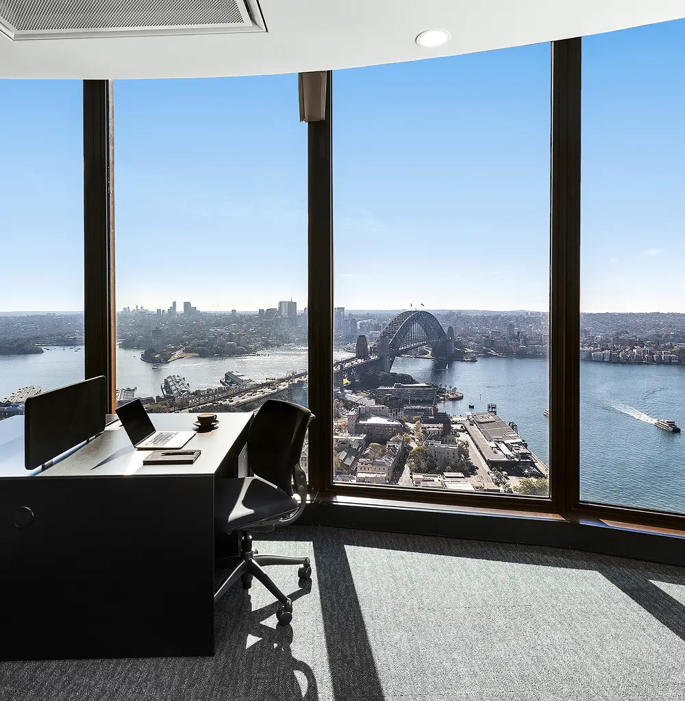 A modern office desk equipped with a laptop and swivel chair sits beside floor-to-ceiling windows, overlooking a panoramic high-rise view of the Sydney Harbour Bridge on a bright, clear day.