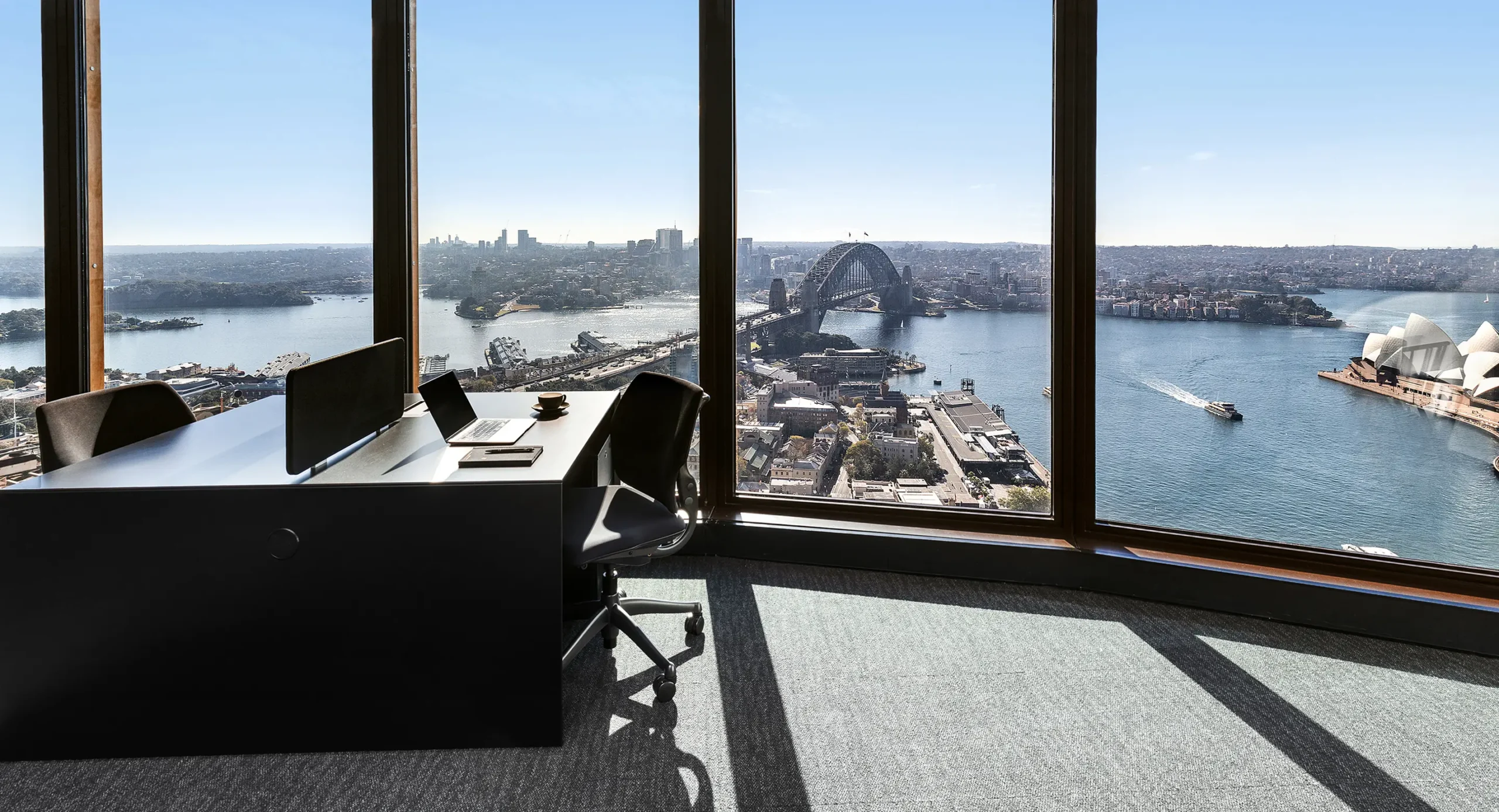 A modern black office desk equipped with a laptop sits beside floor-to-ceiling windows, offering panoramic views of Sydney Harbour, the Harbour Bridge, and the Sydney Opera House.