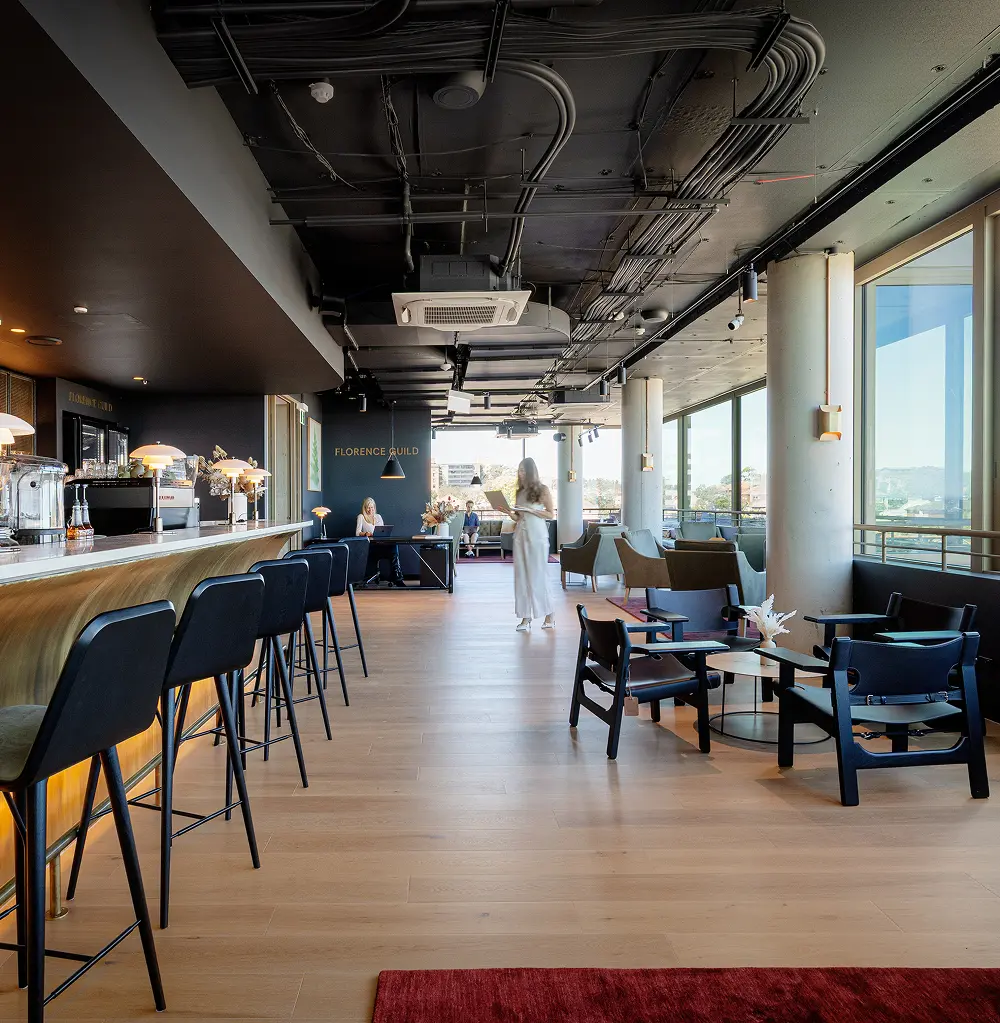 A woman in white walks through a modern co-working lounge holding a device, surrounded by a bar with black stools, armchairs, and exposed ceiling cables. Text reads "FLORENCE GUILD".