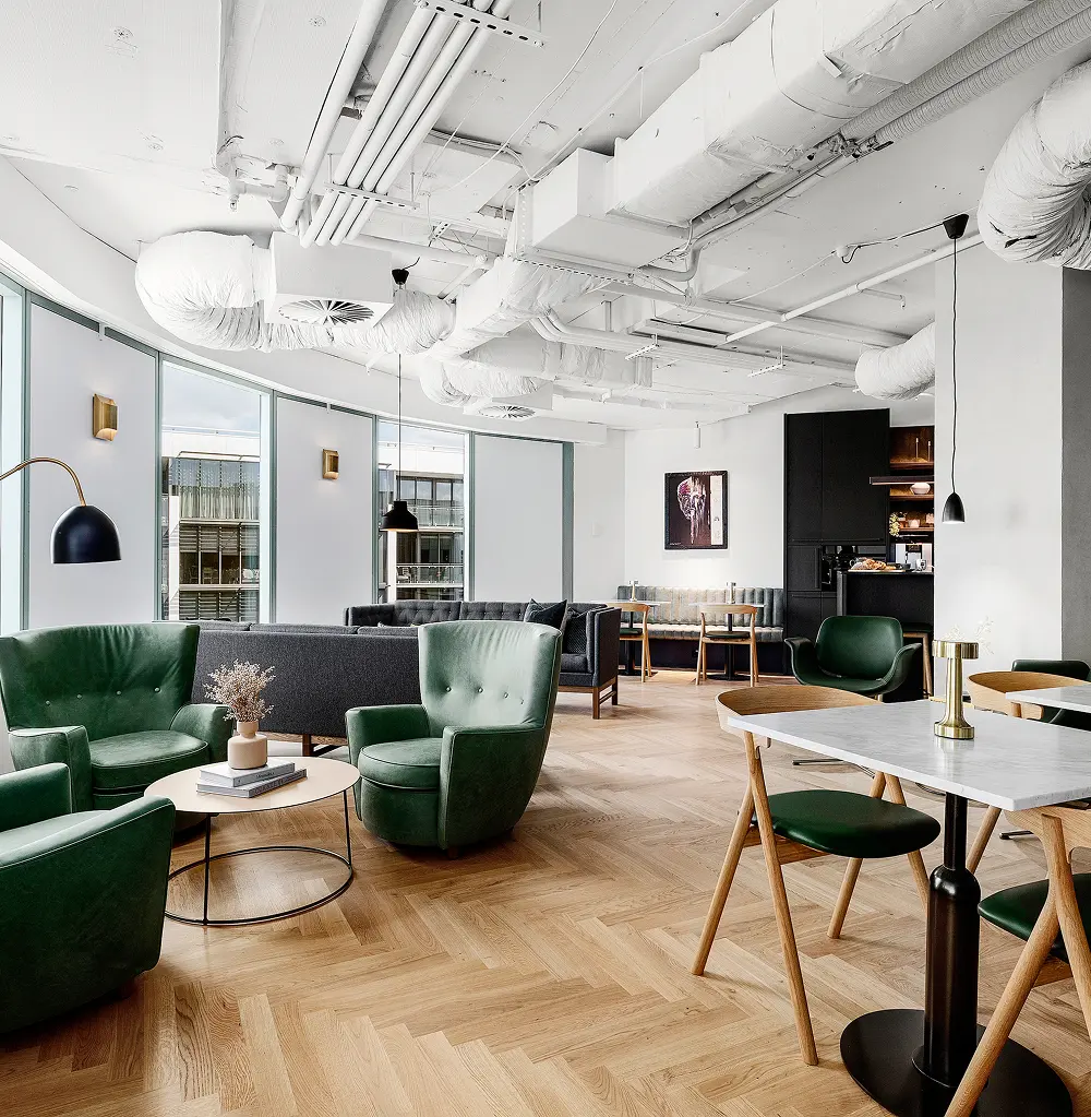 Plush green leather armchairs and sleek sofas are arranged throughout a modern lounge featuring light herringbone wood floors and an exposed white ceiling with industrial ductwork.
