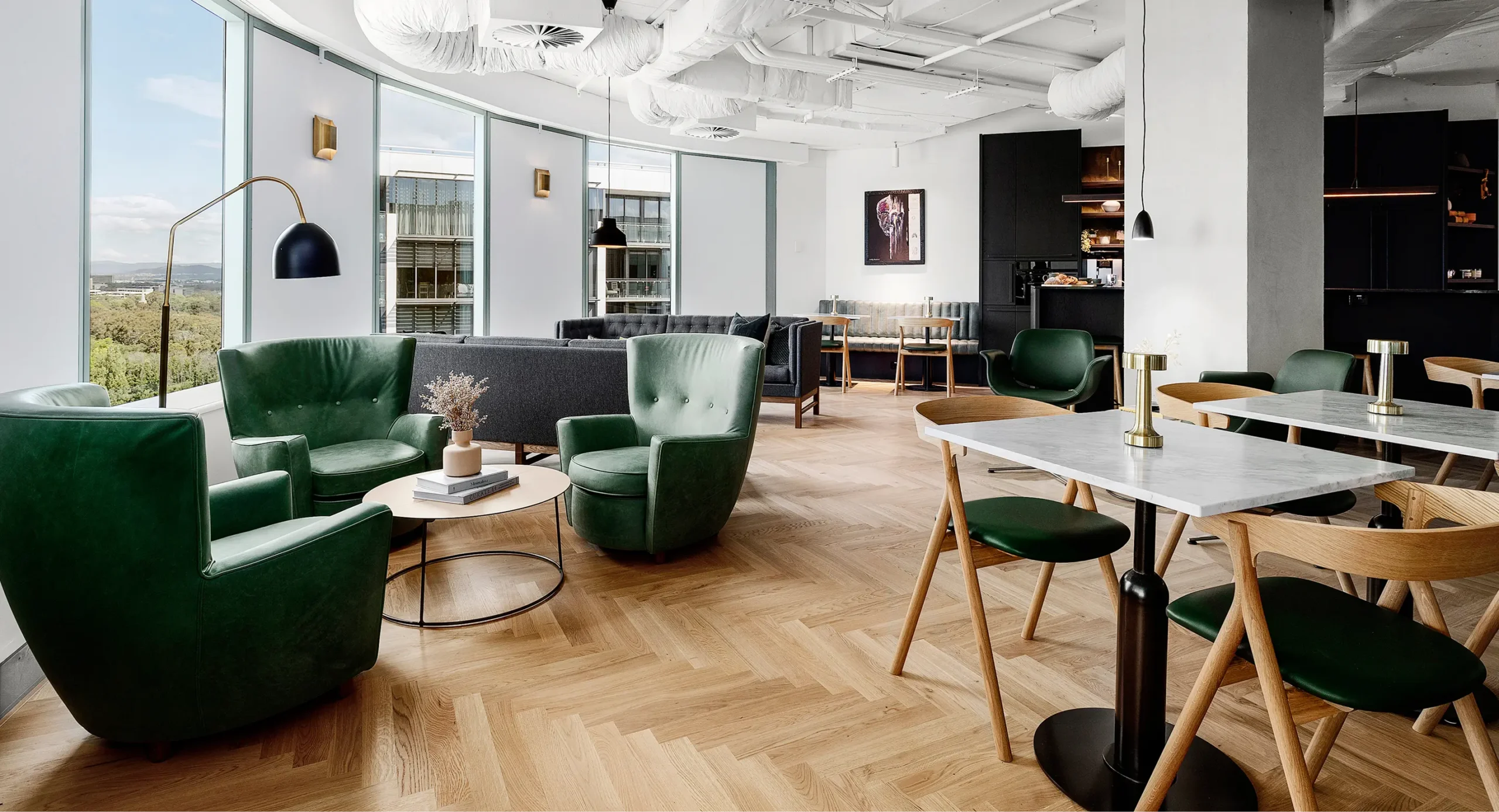 Green leather armchairs and marble-topped dining tables fill a modern lounge space featuring light-wood herringbone flooring, large floor-to-ceiling windows with city views, and exposed white ceiling ducts.