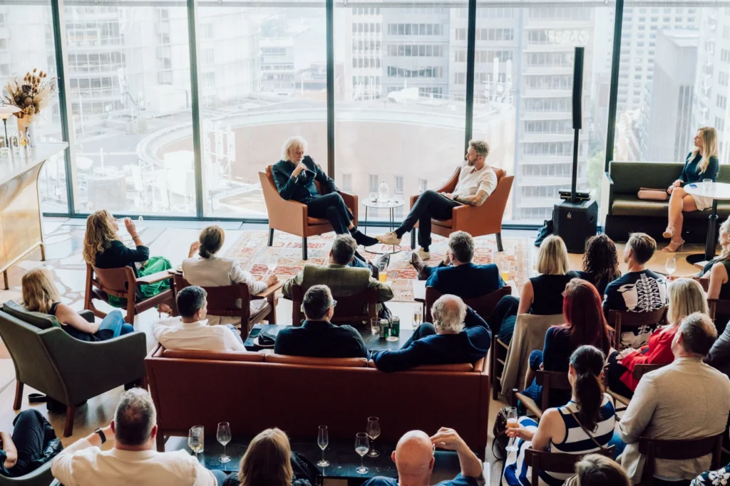 Two speakers sit in armchairs conducting a live talk before a seated audience, inside a high-rise room with floor-to-ceiling windows framing city skyscrapers.