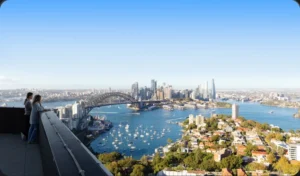Two women lean over a balcony railing, looking at a panoramic view of Sydney Harbour, the Harbour Bridge, and distant city skyscrapers on a bright, clear day.
