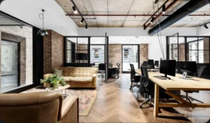 An empty modern office features a leather sofa lounge area alongside long wooden desks with computer monitors, set on herringbone wood floors beneath exposed concrete ceilings and brick walls.