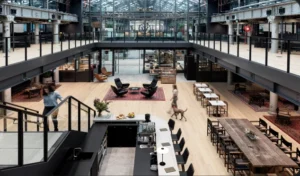 People walk through a vast, modern indoor atrium featuring light wood flooring, a coffee bar, lounge and dining areas, and a black-framed mezzanine level overhead.