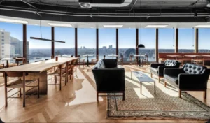 Black leather sofas and a long wooden table sit in a spacious, sunlit office lounge featuring floor-to-ceiling windows that offer panoramic views of a city skyline and harbor.