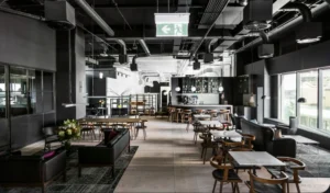 A modern, industrial-style lounge features black leather sofas, wooden dining tables, and a bar counter, set across light wood floors beneath a dark exposed ceiling with ventilation pipes.