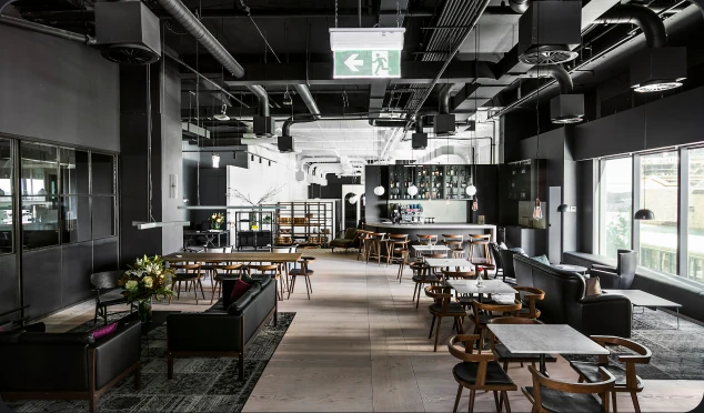A modern, industrial-style lounge features black leather sofas, wooden dining tables, and a bar counter, set across light wood floors beneath a dark exposed ceiling with ventilation pipes.