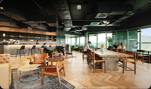 People sit and converse in a spacious, modern lounge featuring leather armchairs, a long wooden communal table, a sleek bar, and large windows revealing an outdoor landscape.
