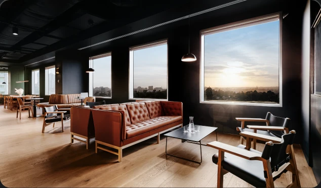 Brown leather tufted sofas and black-seated armchairs frame a sleek coffee table inside a modern, dark-walled lounge featuring light wooden floors and large windows overlooking a city at sunset.