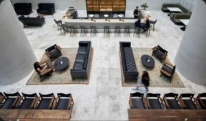 Patrons relax, work on laptops, and sit at a long bar inside a spacious, tiled atrium lounge furnished with leather sofas, patterned rugs, and large concrete columns.