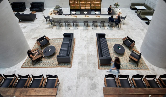 Patrons relax, work on laptops, and sit at a long bar inside a spacious, tiled atrium lounge furnished with leather sofas, patterned rugs, and large concrete columns.