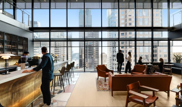 People mingle and order drinks inside a modern, spacious high-rise lounge equipped with leather seating, featuring floor-to-ceiling windows that overlook a skyline of city skyscrapers.