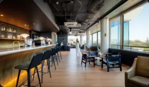 A woman in white walks through a modern lounge featuring a long brass bar with tall stools, dark seating, and floor-to-ceiling windows overlooking trees. Text on the wall reads "FLORENCE GUILD".